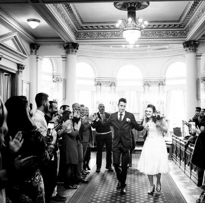 Photo en noir et blanc des mariés sortant de la salle de mariages commune d'Ixelles en dansant lors de leur mariage
