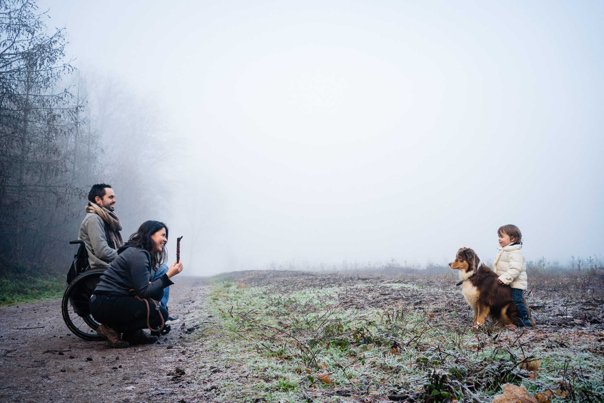 Maman est accroupie sur un chemin de campagne avec un baton dans les mains devant sa fille et son chien lors d'un reportage photo en famille.