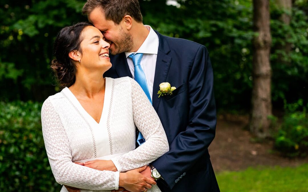 Une mariée adossée à son mari avec ses mains autour de sa taille dans un jardin de Rixensart, Brabant Wallon.Travailler avec des photographes de mariage expérimentés