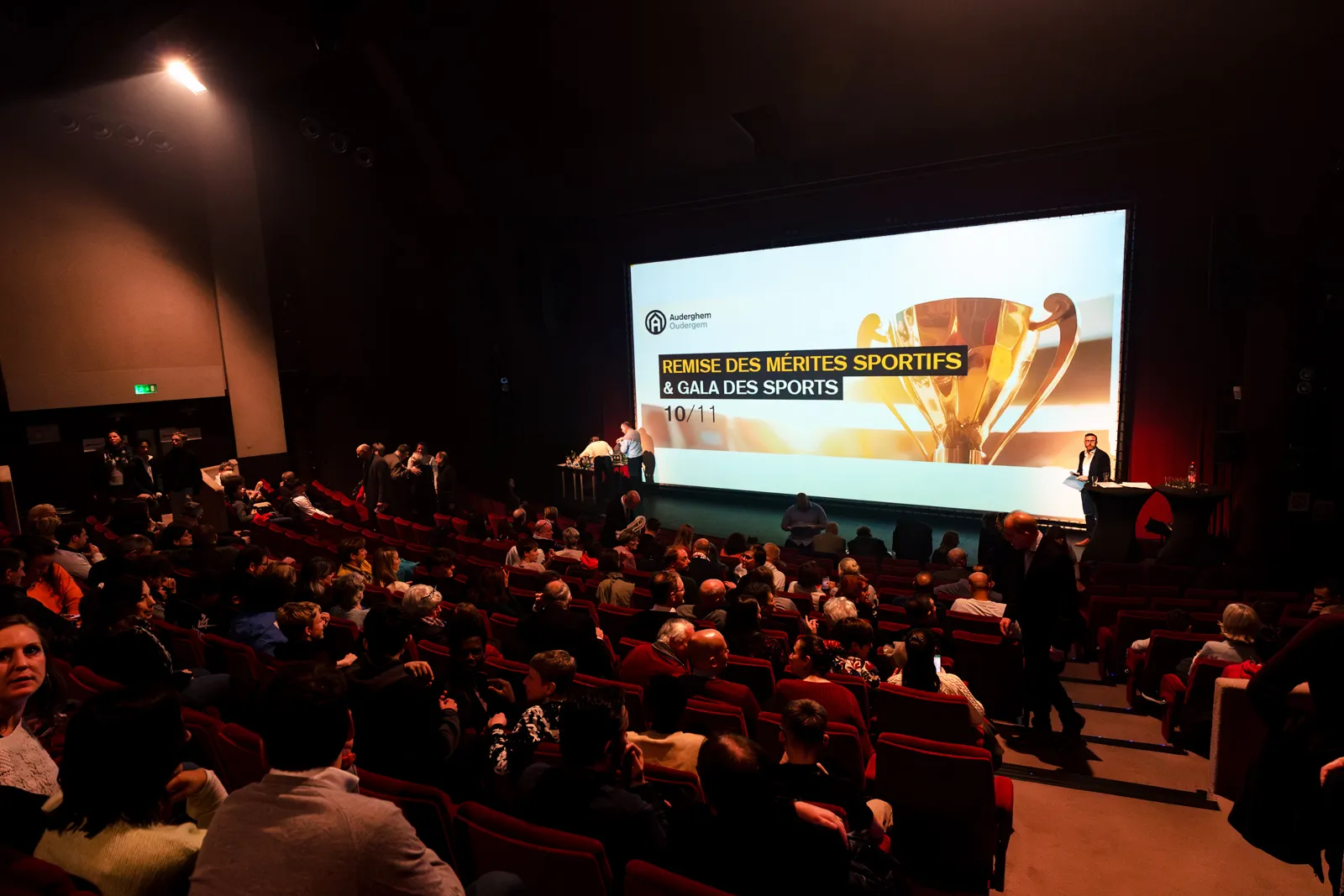 Vue d'une salle de spectacle à Auderghem, Bruxelles avec un écran géant sur lequel est projeté le thème de la soirée, les mérites sportifs