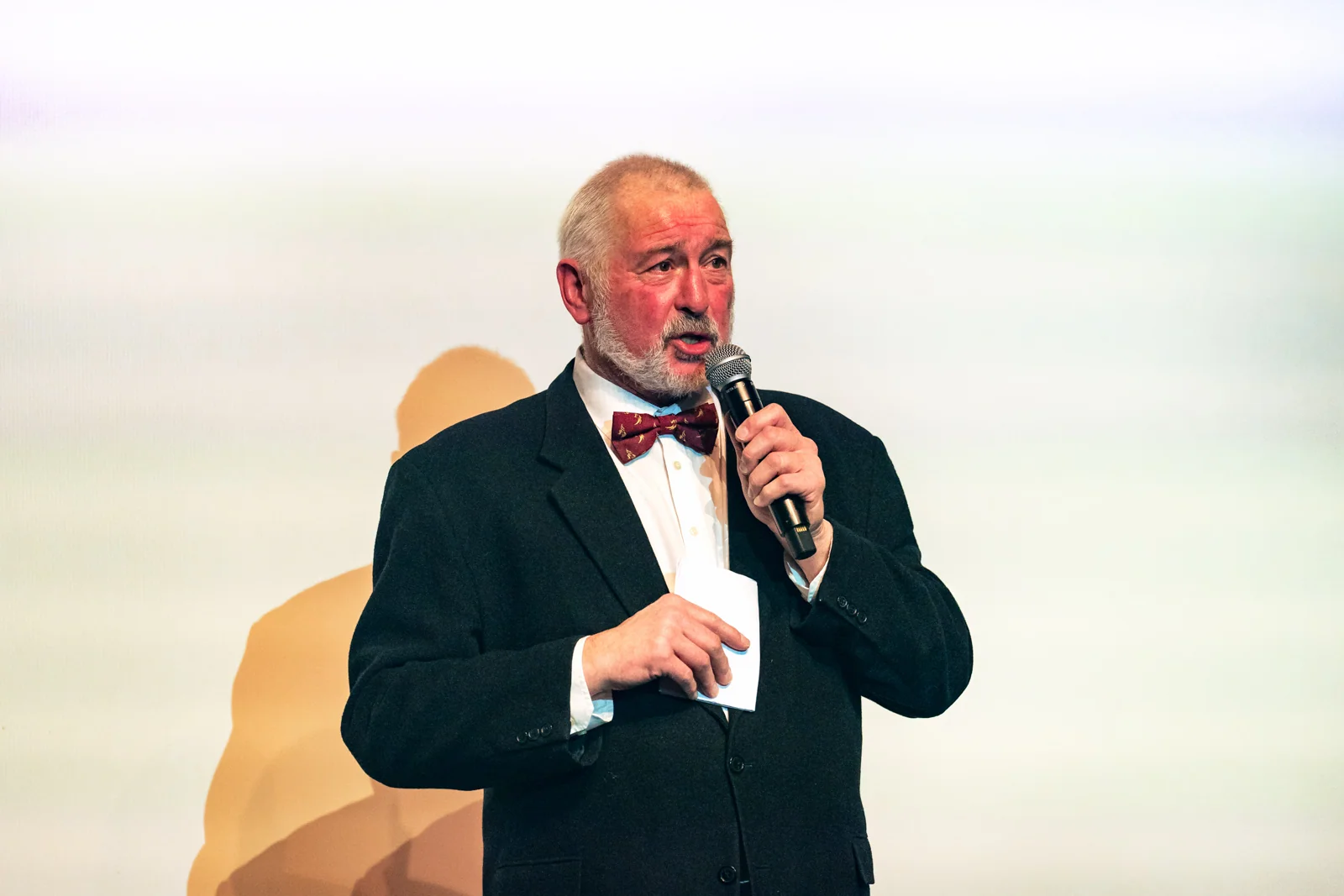 Homme barbu en costume et noeud paillon faisant un discours devant écran géant blanc à Bruxelles.