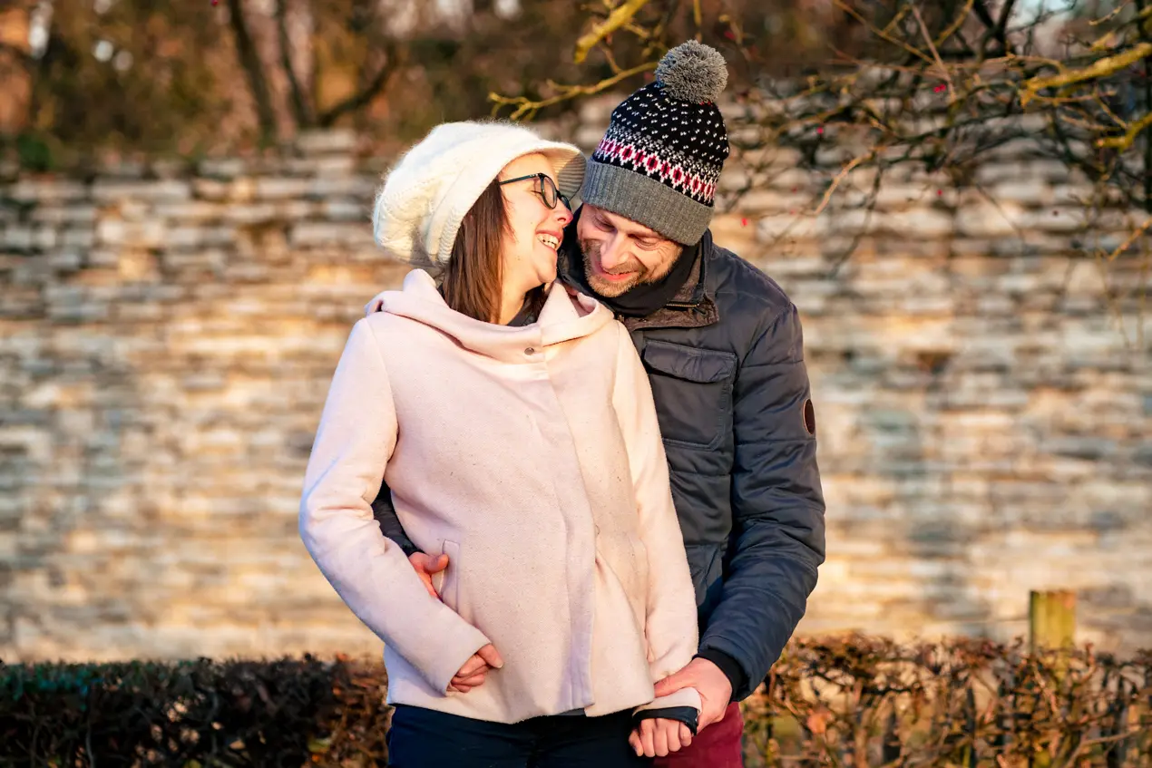 Un homme en bonnet se tient derrière une femme qui lui parle dans l'oreille au soleil en hiver au Rouge Cloitre à Bruxelles