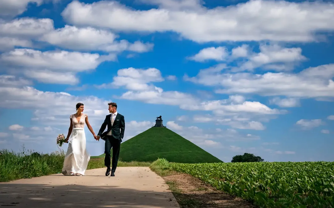 Un couple marché le long d'un champs avec le Lion de Waterloo dans le fond, par un photographe de mariage à Waterloo